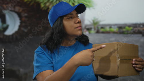 Photos Delivery woman in blue cap and shirt holds a cardboard box and points finger at viewer on street corner; rejection