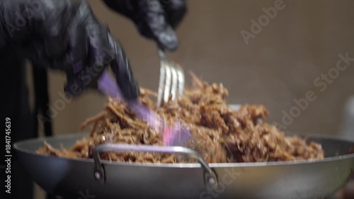 Close-up of juicy shredded pulled meat being prepared with forks in a large pan. Slow cooked barbecue style comfort food with rich texture and savory appearance.