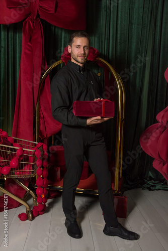 A young handsome man in a black shirt holds a gift and stands near a luggage cart in a hotel interior