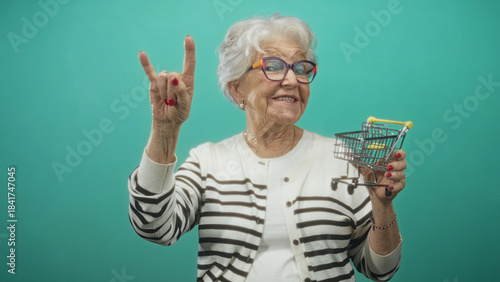 Quadro su tela Woman wearing striped cardigan and eyeglasses holding a miniature shopping cart and flashing rock hand sign in studio; playful joy