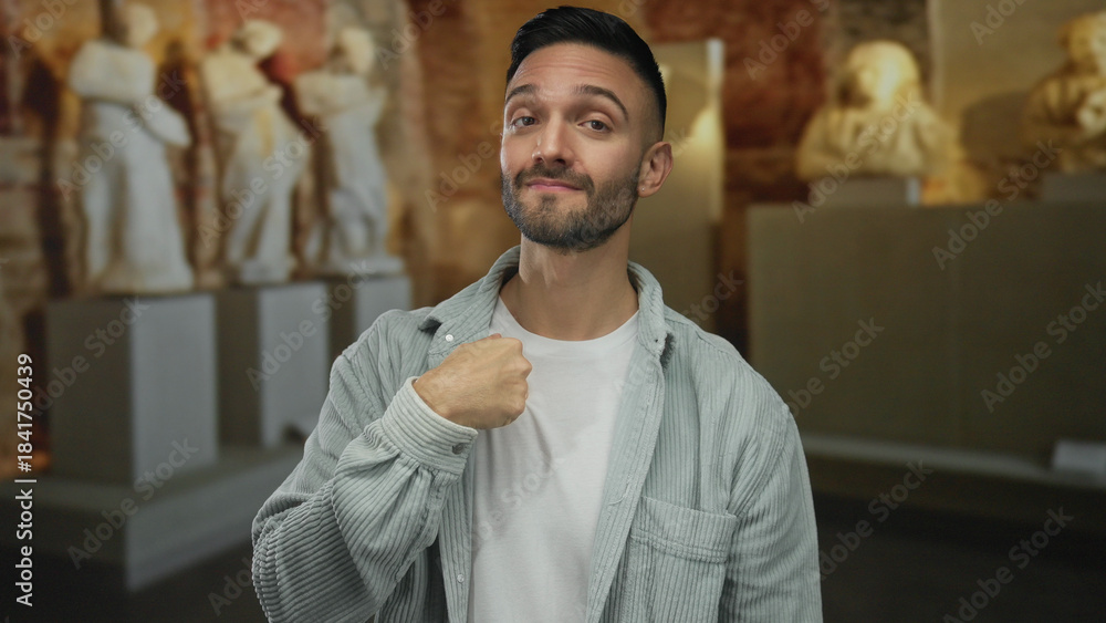Obraz premium Hispanic man in an art gallery pointing at himself, surrounded by sculptures in an indoor setting, suggesting self-reference or identity exploration.