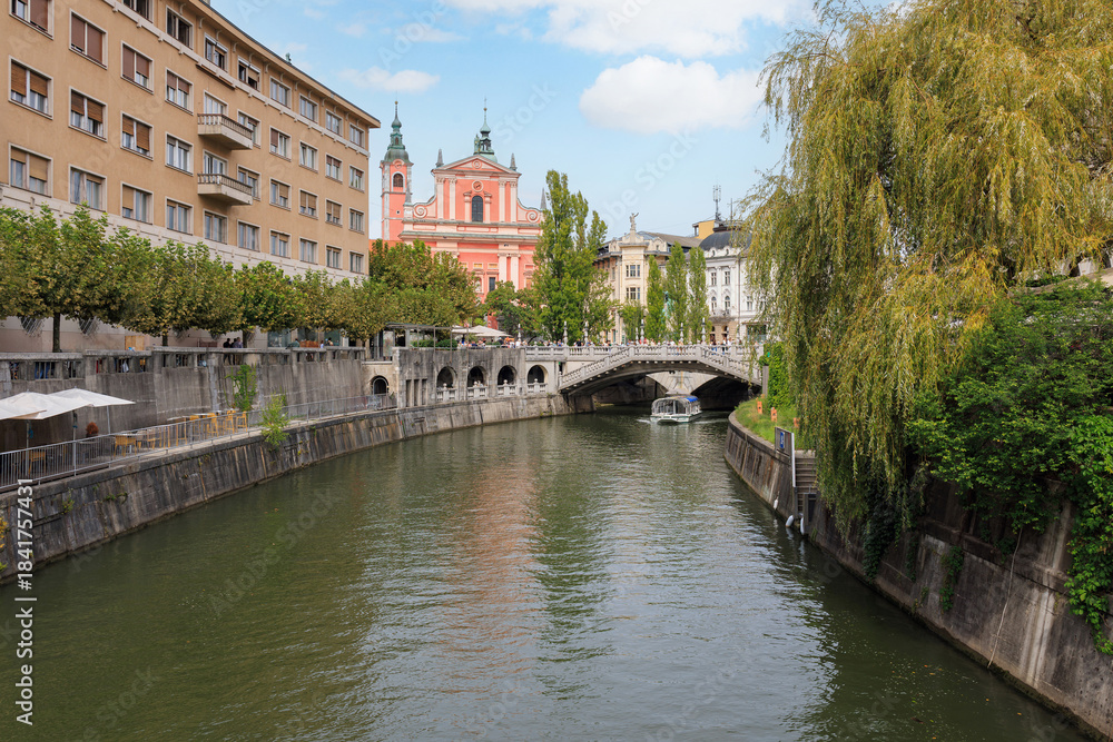 Fototapeta premium People stroll near the Ljubljanica River, showcasing the beautiful architecture and trees in Ljubljana on a sunny day.