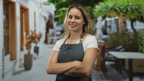 Young blonde woman with crossed arms wearing denim apron and white tee smiling at a street cafe terrace during daytime; confidence. Free Download Young blonde woman with crossed arms wearing denim apron and white tee smiling at a street cafe terrace during daytime; confidence. - GFXLoad