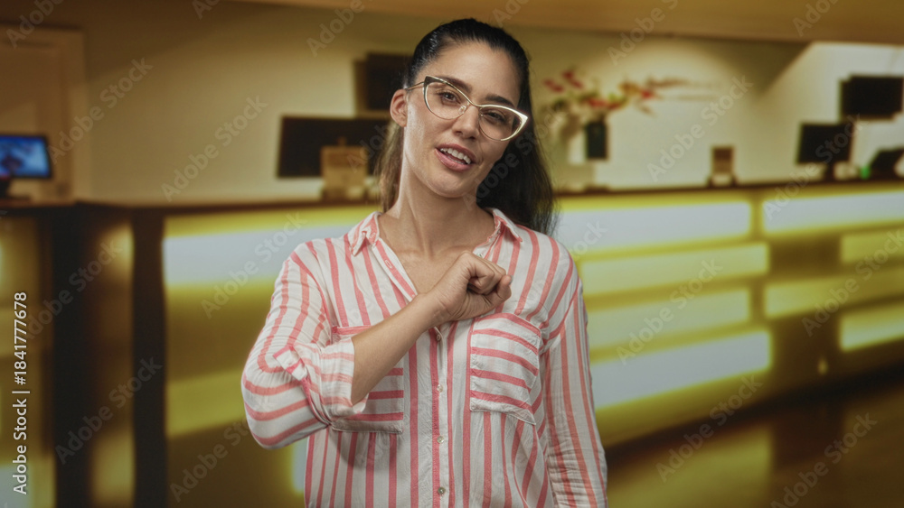 Obraz premium Woman with fist to chest gesture at hotel reception desk inside a building, wearing pink striped shirt and glasses; solidarity.