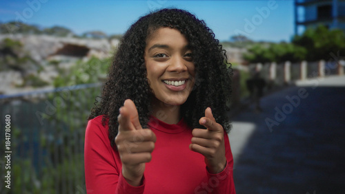 Woman in red shirt points finger to camera beside fence on paved walkway in sunlit park; playfulness.