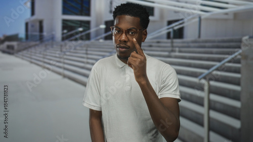 Man wearing white polo shirt pointing finger by modern building staircase while glancing sideways; curiosity.