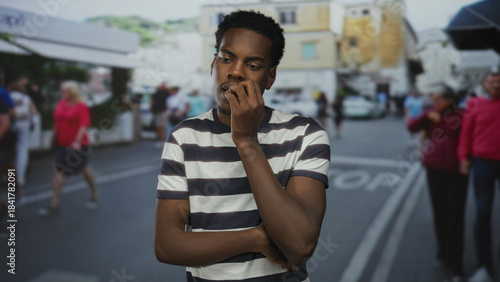 Young african american man in striped shirt biting nails on a busy urban street amid blurred pedestrians; anxiety.