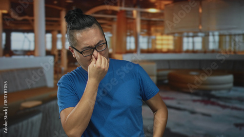 Bild auf Leinwand Man pinches nose with fingers in restaurant building wearing blue shirt and glasses; disgust smell reaction
