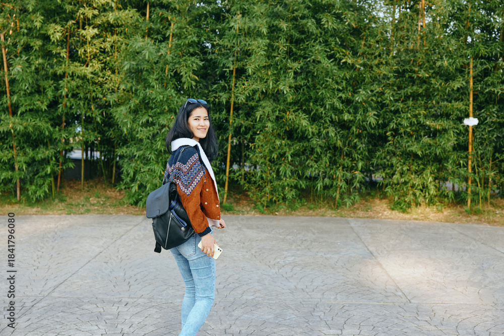 Naklejka premium Woman with backpack walking on a paved path beside green trees, casual style, cheerful pose, outdoor lifestyle and travel vibe for everyday moments