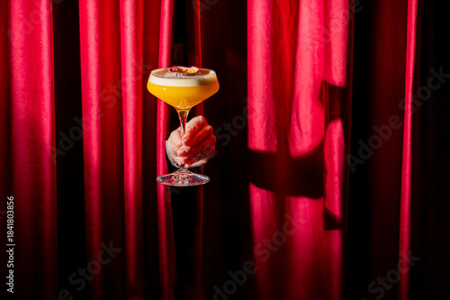 A vibrant yellow cocktail in a coupe glass held by a hand against a luxurious, deep red velvet stage curtain backdrop, with dramatic lighting.