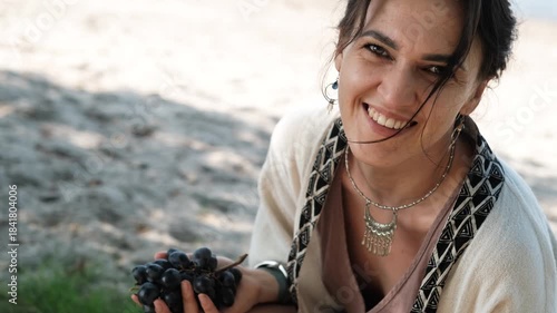 A beautiful brunette in a bohemian style relaxes outdoors in summer against a backdrop of a green forest, holding a large bunch of juicy black grapes