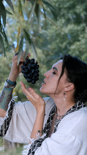 A beautiful brunette in a bohemian style relaxes outdoors in summer against a backdrop of a green forest, holding a large bunch of juicy black grapes