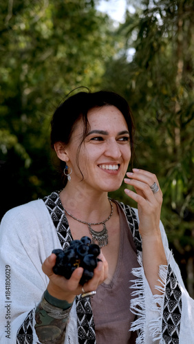 A beautiful brunette in a bohemian style relaxes outdoors in summer against a backdrop of a green forest, holding a large bunch of juicy black grapes