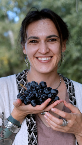 A beautiful brunette in a bohemian style relaxes outdoors in summer against a backdrop of a green forest, holding a large bunch of juicy black grapes