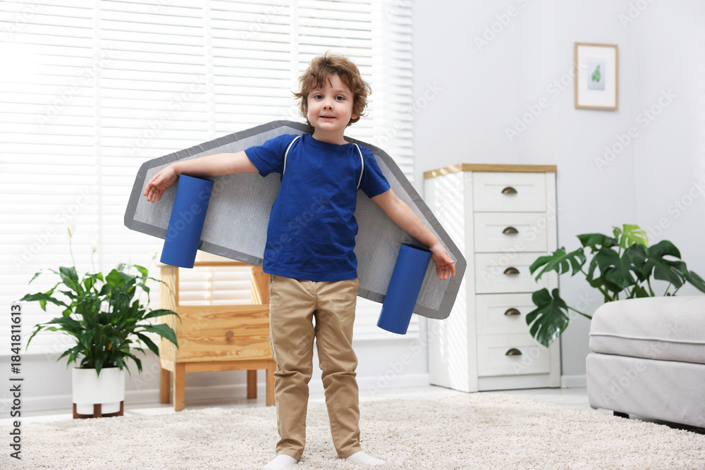 Fototapeta premium Little boy with cardboard plane wings playing pilot at home