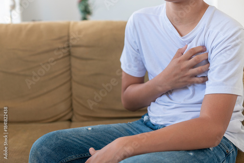 A young man sits on a couch with his hand on his chest. He is wearing a white shirt and blue jeans