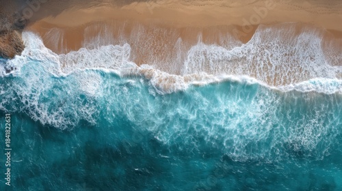 Waves break on the shore splashing white foam against the sand. The ocean water appears clear blue with a bright sun high in the sky.