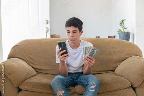 A young man sits on a couch with a cell phone in his hand and a pile of cash next to him. He is checking his phone while looking at the money
