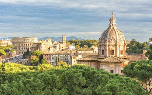 Panoramic view of Rome with the dome of Santissimo Nome di Maria al Foro Traiano and the Colosseum in the background, bathed in warm sunlight above lush green trees.