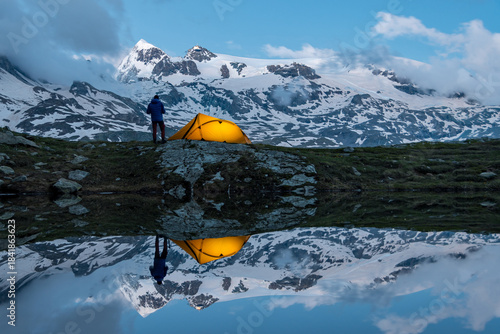 Trekker accanto a tenda illiminata riflessi sul lago, con Monte Rosa sullo sfondo, campeggio selvaggio (wild camping), valtournenche, Italia