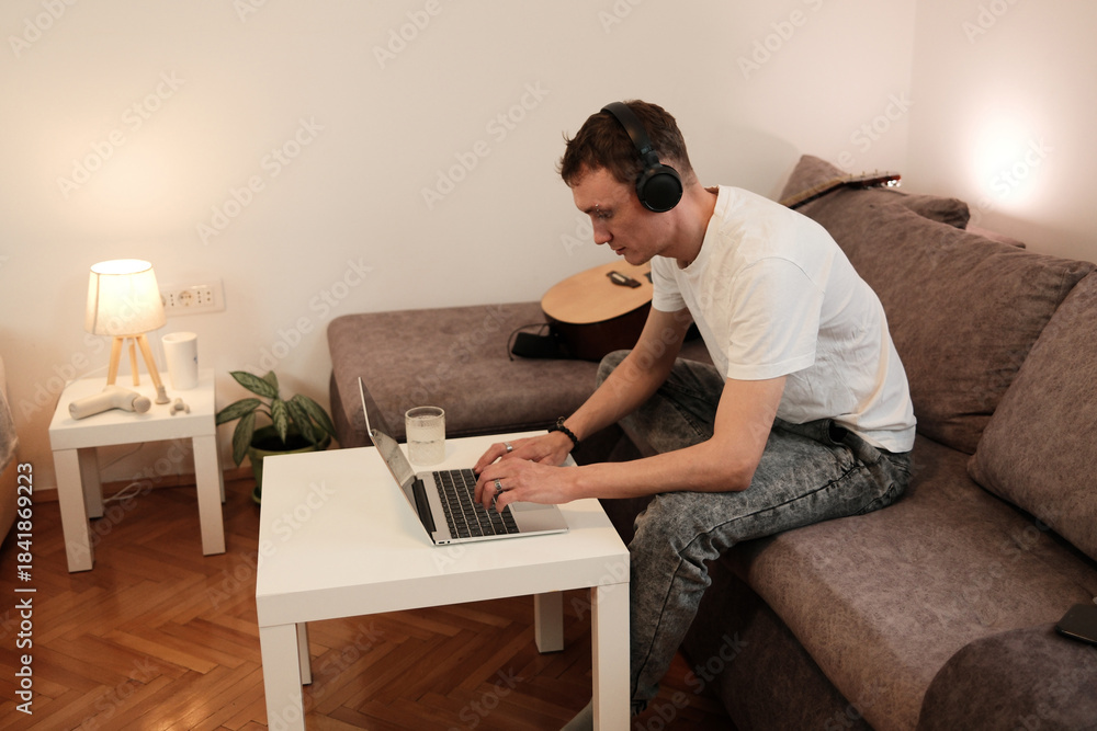 Fototapeta premium A man works on his laptop while sitting on the sofa. The concept reflects productivity and remote work in a relaxed home environment.