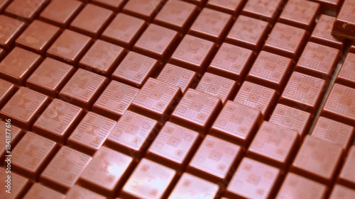 A close-up, high-angle shot captures hundreds of small, unwrapped milk chocolate bars arranged in neat, repetitive rows, ready for the next stage of food production and packaging