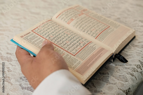 A close-up of a hand reading the Quran on a lace-covered table. Clear Arabic text, warm lighting, and peaceful atmosphere suitable for themes of Islam, Ramadan, prayer, study, and spirituality.