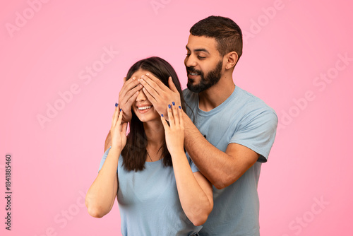 A young couple shares a fun and affectionate moment as the man playfully covers the woman's eyes. They are both smiling and wearing matching blue shirts, set against a vibrant pink backdrop.