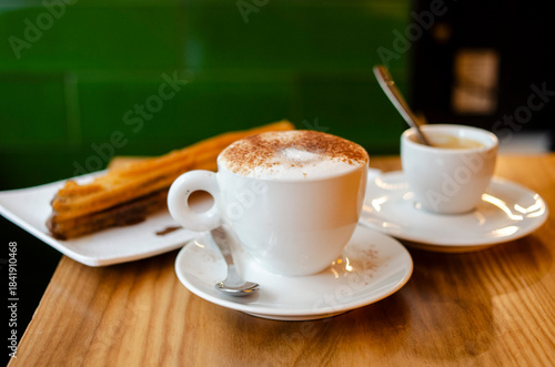 A trio of Spanish cafe treats -- a warm, frothy coffee with cinnamon, an espresso and a churro with chocolate on a wood table. Horizontal image.
