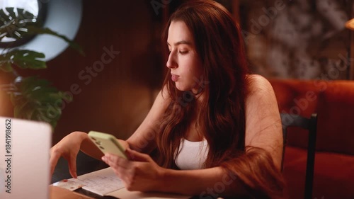 Beautiful woman at desk checking phone while studying late evening with laptop and notes, focused expression, warm lamp light, cozy apartment study scene showing concentration and quiet productivity