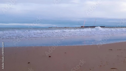 Tranquil seaside landscape with stormy coastal surf water waves rolling on empty sand beach on Baltic sea coast and seaport quayside on background at cloudy autumn day. Low angle shot seascape.