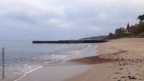 Tranquil seaside landscape with calm coastal surf water waves rolling on sand beach and old wood breakwater of Baltic sea shore under cloudy sky at overcast autumn morning or dusk. Low angle shot.