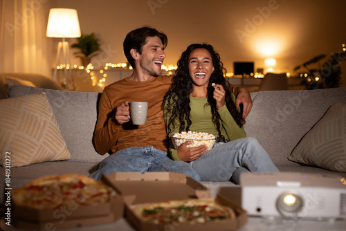 Cuadro en lienzo Cheerful couple relaxes on sofa, laughing while watching a comedy movie with a home cinema projector