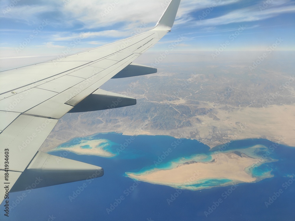 Obraz premium Aerial View of Airplane Wing Over Coastline and Sea, Red Sea, Egypt - A high-resolution aerial photograph taken from an airplane window, showing the aircraft wing above a deep blue sea with a desert c