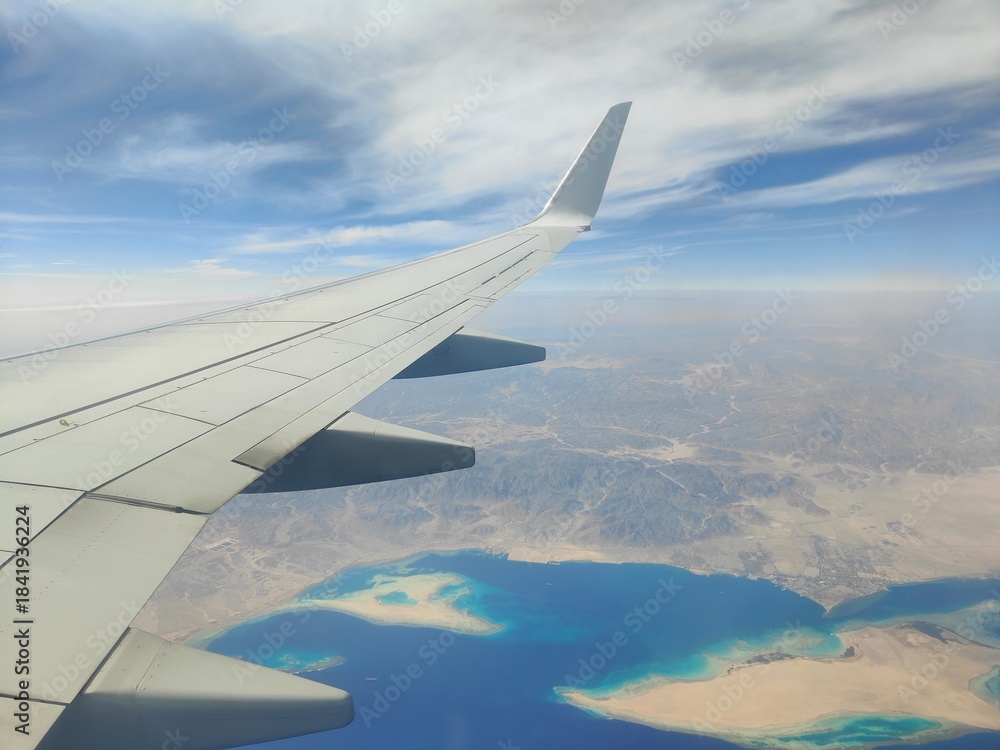 Obraz premium Aerial View of Airplane Wing Over Coastline and Sea, Red Sea, Egypt - A high-resolution aerial photograph taken from an airplane window, showing the aircraft wing above a deep blue sea with a desert c