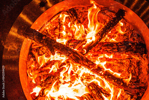 Intense flames engulf logs in an outdoor fire pit at night. The fire pit is circular with charred wood arranged in a cross pattern, surrounded by glowing embers warm orange and yellow hues and steel 