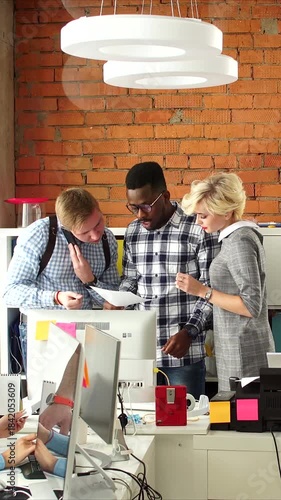 vertical video of team collaborating around desktop computer in modern brick office, designer points to screen while developer and project manager review documents