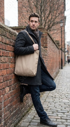 Stylish man in black coat and jeans leaning against a brick wall with a tote bag.