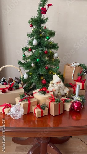 Merry Christmas and Happy New Year! Vertical video of Christmas tree decorated with colorful balls, garlands, and pile of gifts in paper boxes with red ribbons on table. Holiday background