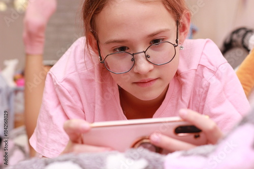 Close-up of a person lying on a bed and holding a smartphone in landscape orientation while playing a mobile game. The scene features casual pink clothing and a cozy bedroom interior with a soft-focus