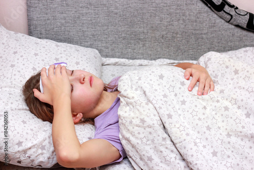 Close-up of a young girl lying in bed with her eyes closed, feeling unwell. She has a cold compress or ice pack on her forehead, indicating a fever or headache. Wrapped in a blanket with a star patter