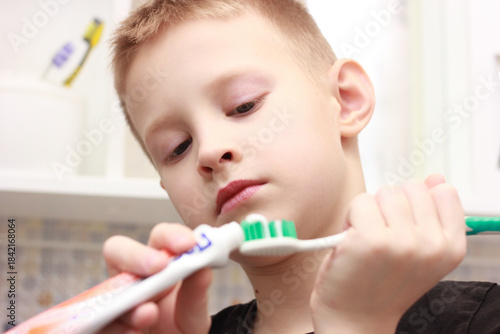 A close-up of a small boy, concentrating on squeezing toothpaste onto a brush. The image symbolizes the importance of daily oral hygiene and teaching children to be independent