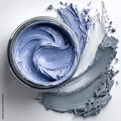 Close up of a jar filled with textured blue cosmetic cream, with grey powder swirled around it against a clean white backdrop.