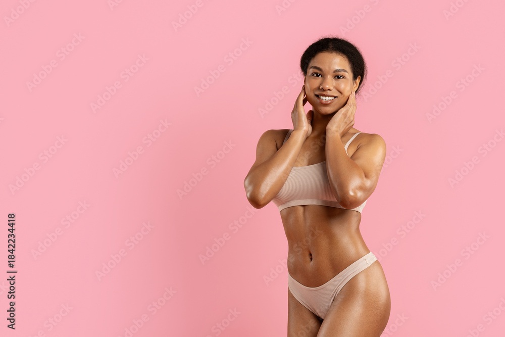 Naklejka premium Young black woman posing playfully in beige underwear on pink studio background. Fit physique, glowing skin and cheerful expression perfect for wellness, skincare and lifestyle visuals