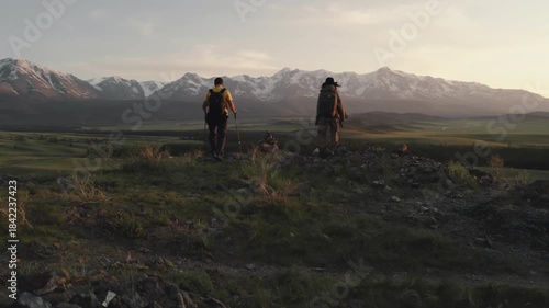 Two young active trekkers approach the edge of a cliff and look at the panorama of the high Altai Mountains at sunset. The spirit of adventure, hiking, summer travel in the wild nature