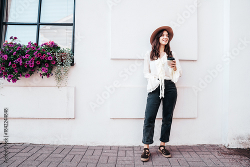 Lifestyle portrait of young stylish woman dressed in white blouse. Model walking with coffee cup in old town in sunny day. Beautiful smiling female in hat. Cheerful and happy. Takeaway coffee