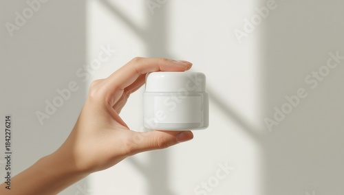 Female hand holding blank white cosmetic jar with moisturizer cream or balm against sunlit background