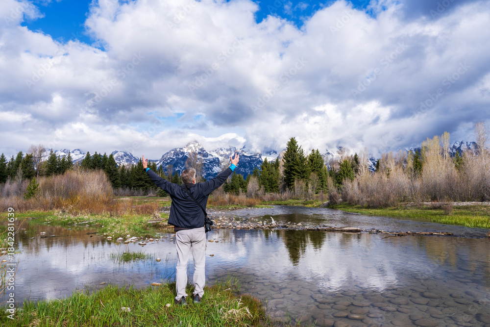 Fototapeta premium At Grand Teton National Park, a traveler lifts an arm, savoring the harmony of sky, peaks, and river reflections.