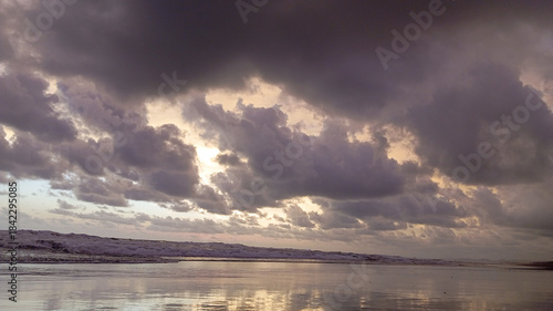 ​A moody coastal scene featuring heavy dark storm clouds over a reflective beach, with crashing white waves under dim light