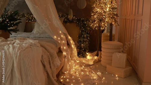 Cozy Christmas at home. New year night.Warm evening in interior bedroom with bed with baldachin, christmas tree, gifts box and illimination many glowing golden lights garland. Basket with branch fir. 